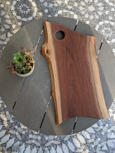 walnut live edge charcuterie board with hole for hanging. Cheese board resting on grey round table with succulents in a blue pot to the side.