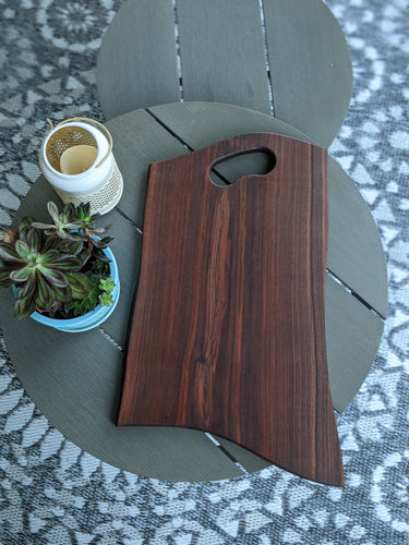 Dark walnut charcuterie board with grip handle on grey table. Green succulants in blue pot to the side.